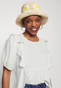 Blusa branca com franzidos e tecido texturizado, com detalhes em rendas ao centro, acompanhada de um chapéu de palha adornado com flores amarelas.