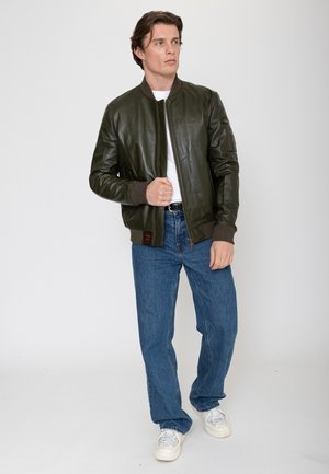 Man die een donkergroene leren bomberjack draagt, wit T-shirt, blauwe spijkerbroek en witte sneakers, staand tegen een effen lichte achtergrond.