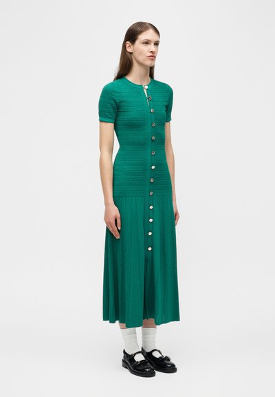 Femme debout portant une robe en maille verte à manches courtes et boutonnée, avec des chaussettes blanches et des mocassins noirs, devant un fond clair uni.