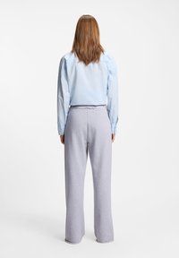 Άτομο με ίσια καστανά μαλλιά που φοράει φωτεινό μπλε πουκάμισο με μακριά μανίκια και άνετο γκρι παντελόνι, στεκόμενο με την πλάτη γυρισμένη σε λευκό φόντο.