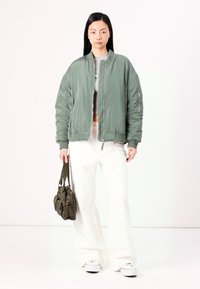 Veste bomber rembourrée dans un vert terne, avec une fermeture éclair sur le devant, des poignets côtelés et des poches latérales. Associée à un pantalon blanc à jambes larges et un petit sac.