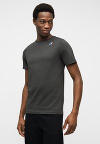 Νεαρός άνδρας που κοιτάζει πλάγια φορώντας σκούρο γκρι t-shirt με κοντά μανίκια και μικρό πολύχρωμο λογότυπο στο στήθος.