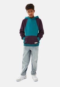 Hoodie med teal- och lila paneler, känguruficka och ribbade muddar. I kombination med ljusblå jeans och vita sneakers.