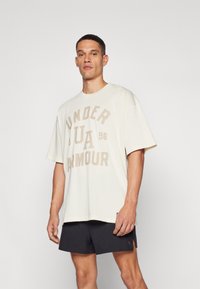 Camiseta oversize color crema con "UNDER ARMOUR" y "UA 96" impresos en beige. Manga corta, escote redondo. Llevada con pantalones cortos negros.
