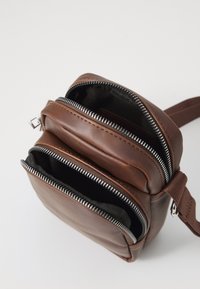 Brown leather bag with two zippered compartments, featuring silver zippers and a textured surface. Includes a brown adjustable strap.