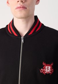 Black bomber jacket with red striped collar, zip front, and embroidered crest detailing, featuring a smooth texture and fitted design.