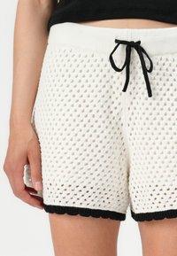 White knitted shorts featuring a textured pattern, black drawstring waistband, and scalloped black trim at the hem.