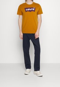 Musztardowożółta bawełniana koszulka z fioletowym logo graficznym Levi's. Połączona z granatowymi spodniami i białymi płóciennymi trampkami.