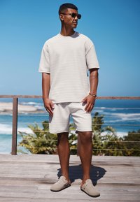 Ljusgrå texturerad kortärmad skjorta, matchande shorts och grå slip-on skor, bärs med runda solglasögon framför en blå havsbakgrund.