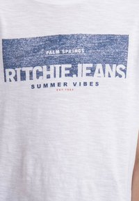 T-shirt en coton blanc avec un texte bleu qui dit « PALM SPRINGS RITCHIE JEANS SUMMER VIBES » et un détail rouge « EST. 1989 ».