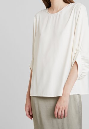 Femme portant un chemisier crème ample à manches longues avec des manches froncées et une jupe gris clair sur un fond uni.