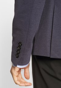Námořnický blazer s texturou na rukávu, který má manžetu se pěti knoflíky a černými knoflíky. Světle modrá podšívka je viditelná na okraji manžety.