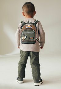Un sac à dos marron et vert avec un logo de soleil souriant, des poches latérales et des détails de porte-clés. Un enfant portant une chemise claire et un pantalon vert, tourné le dos.