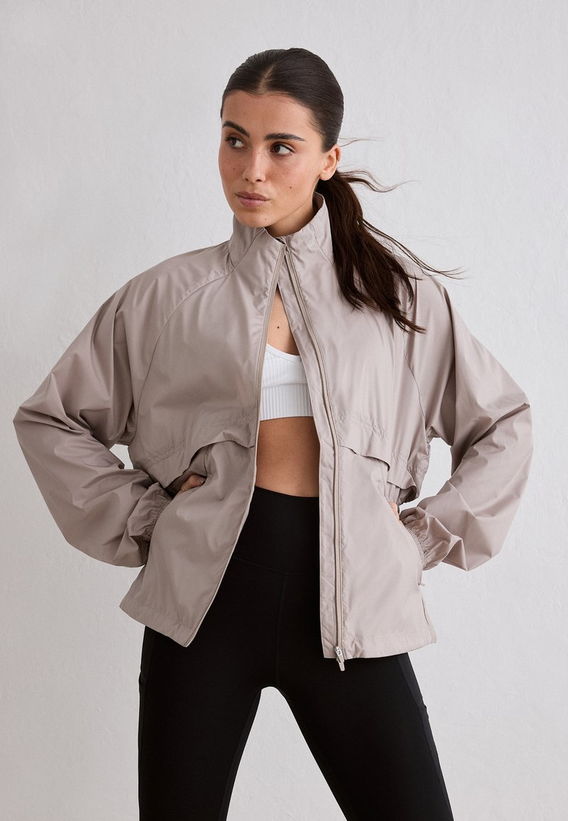 Femme aux cheveux foncés attachés en queue de cheval, portant un coupe-vent beige zippé par-dessus un brassière de sport blanche et un legging noir, debout les mains sur les hanches.