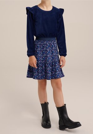 Marineblauw fluwelen top met lange mouwen en ruches; gecombineerd met een wijd uitlopende marineblauwe rok met kleurrijke bloemmotieven. Zwarte enkellaarsjes.