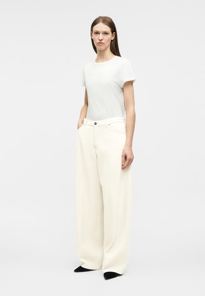 Femme portant une chemise blanche à manches courtes et un pantalon large couleur crème avec des chaussures noires pointues, debout les mains dans les poches devant un fond uni.