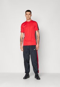 Homme debout portant des vêtements de sport Nike rouges et noirs avec des tatouages sur les deux avant-bras contre un fond clair uni.