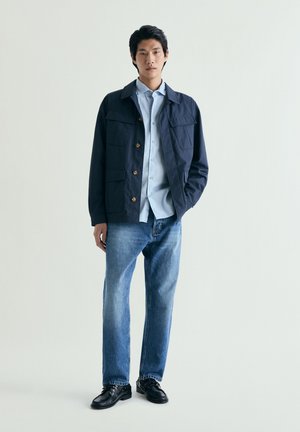 Hombre con chaqueta azul marino sobre camisa de botones azul claro, vaqueros azules y zapatos negros, de pie con una mano en el bolsillo.