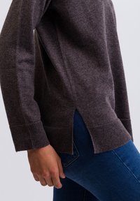 Mörkbrun stickad tröja med ribbade ärmslut och sidofickor, i kombination med blå jeans. Mjuk textur och avslappnad passform synlig.