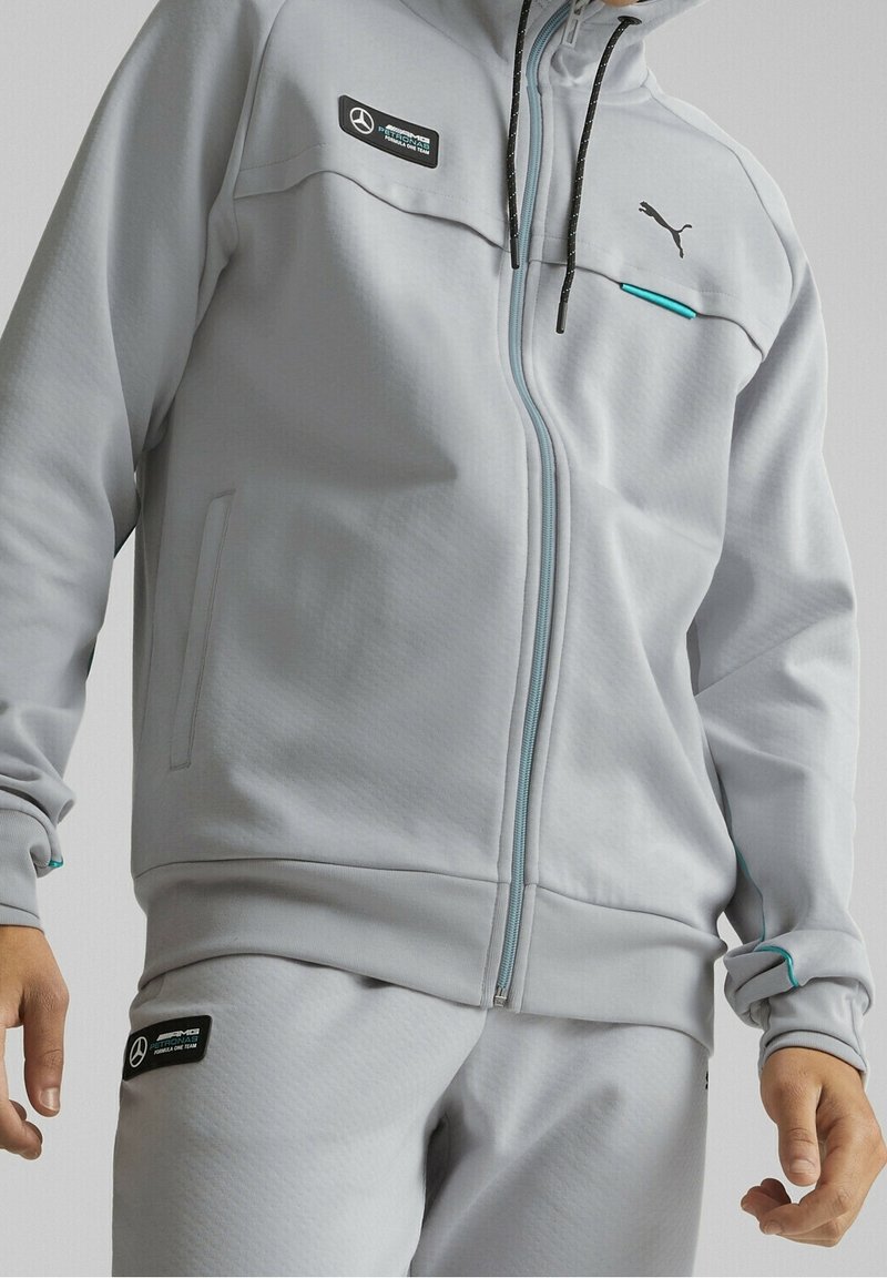 Chaqueta gris con cremallera hecha de tela texturizada. Presenta detalles en negro y azul verdoso, parches de logo y capucha con cordón y puntas contrastantes.
