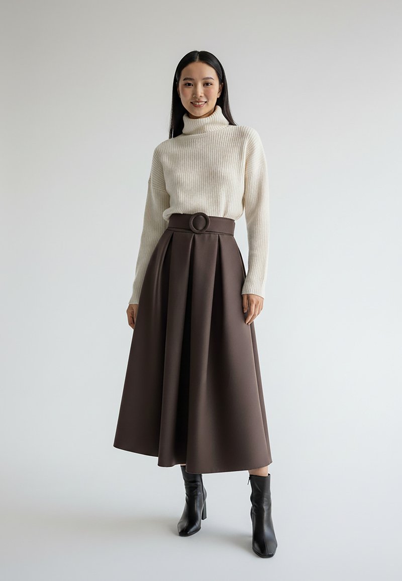 Kremowy prążkowany golf w połączeniu z brązową, plisowaną spódnicą midi z ozdobnym okrągłym paskiem, noszony z czarnymi botkami na obcasie.