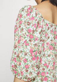 Tissu blanc avec un motif floral rose, pêche et brun sur un vêtement à manches bouffantes, montrant une vue partielle de l'épaule et du cou d'une personne.