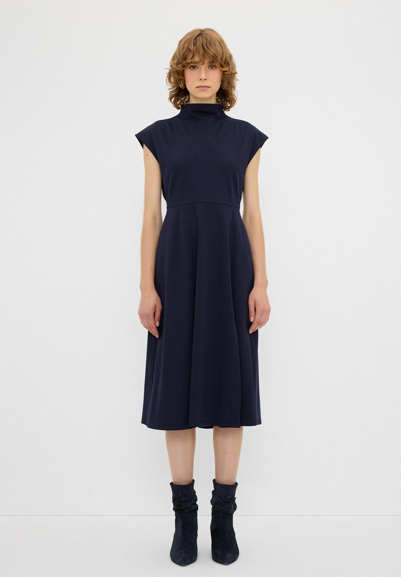 Navy blue, polkuun ulottuva mekko korkealla kauluksella ja hihattomilla hihoilla. Tasainen pinta, tiukka vartalo-osa, levenevä hame. Yhdistetty tummiin, löysiin nilkkasaappaisiin.