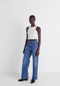 White fitted tank top paired with high-waisted blue straight-leg jeans. Model stands on a neutral background, wearing black boots.