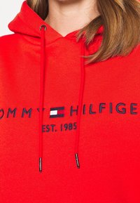 Felpa rossa in misto cotone, con logo blu navy e testo "TOMMY HILFIGER" e "EST. 1985", dotata di coulisse regolabile con punte metalliche.