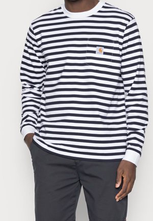 Męski model ubrany w czarno-białą koszulę w poziome paski z długim rękawem i małym logo na kieszeni, połączoną z ciemnymi spodniami.