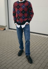 Pull en argyle rouge, noir et bleu avec des bordures noires, superposé sur une chemise blanche à col; jean bleu; chaussures noires avec semelles texturées.