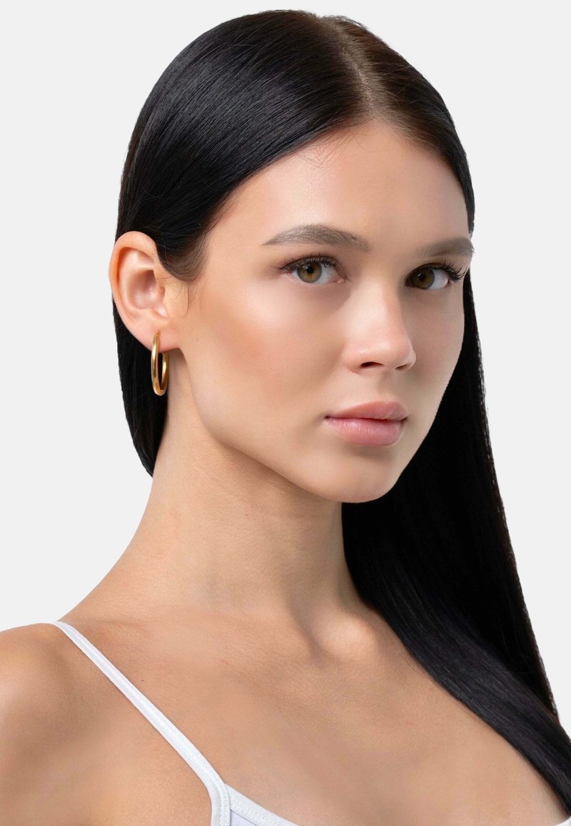 Kuldne rõngaskõrvarõngas modellil, kellel on pikk, sirge must juuksed. Modellil on sile jume ja ta kannab valget varrukateta topi.