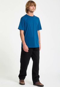 Kortärmad, blå t-shirt i bomull med rund hals, kombinerad med svarta byxor. Framträdande detaljer inkluderar en diskret logga på bröstet.