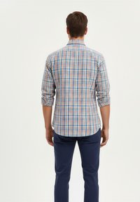 Hombre de pie de espaldas, vistiendo una camisa de manga larga a cuadros en naranja, azul y blanco con las mangas arremangadas y pantalones azul oscuro.