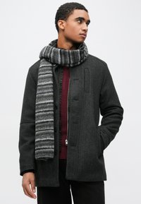 Temno sivi volneni plašč z gumbi spredaj, velikim ovratnikom in bočnimi žepi. Združen z ruto iz sive in črne črtaste pletenine.