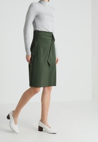 Grüne Wickelrock mit hoher Taille, seitlichem Bindeband und strukturiertem Stoff. Kombiniert mit einem grauen Rollkragenpullover und weißen High Heels.