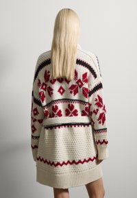 Cárdigan de punto con un corte holgado, que presenta un patrón floral en rojo sobre un fondo crema, con detalles en negro y un diseño de punto texturizado.