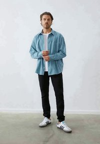 Randig blå och vit skjorta med knapp, bärs över en vit t-shirt, i kombination med svarta byxor och grå sneakers med blå accenter.