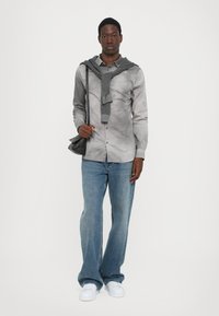 Grå skjorte med knapper og et subtile mønster, parret med brede blå jeans og hvite sneakers; et rutet skjerf er lagt over skuldrene.