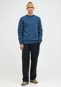 Felpa blu con polsini e scollatura a coste, abbinata a pantaloni cargo neri con tasche con zip e scarpe da trekking beige. Berretto a righe marrone.