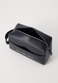 Crna sintetička torbica za toaletne potrepštine sa zaobljenim oblikom, koja sadrži srebrni patentni zatvarač i utisnuti logo "Calvin Klein Jeans" na prednjoj strani.