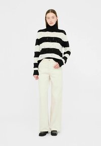 Črno-bela pulover s črtami in visokim ovratnikom, kombiniran z svetlo bež širokimi hlačami in črnimi čevlji. Gladek tekstura in sproščen kroj.