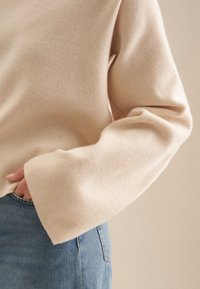 Maglione beige a maglia con maniche a campana, texture liscia, vestibilità rilassata. Abbinato a jeans di denim azzurro, che mostrano un layering informale.