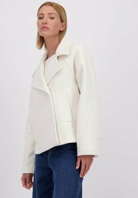 Blonde vrouw in een witte wollen jas met brede revers en zijzakken, gecombineerd met blauwe denim jeans, tegen een effen achtergrond.