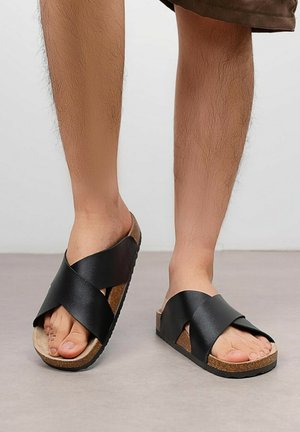 Pieds portant des sandales noires à lanières croisées avec semelles en liège sur un sol gris, personne debout en short marron.