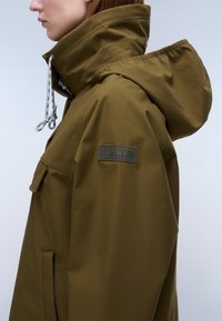 Sideview af en person iført en olivengrøn hættejakke med høj krave og Napapijri-logo på ærmet.