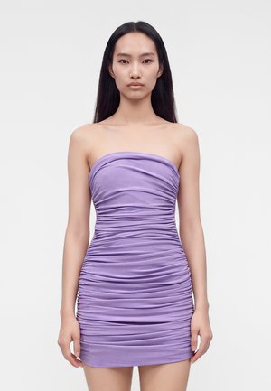 Femme aux longs cheveux foncés portant une mini-robe lilas sans bretelles et froncée, debout devant un fond clair uni.
