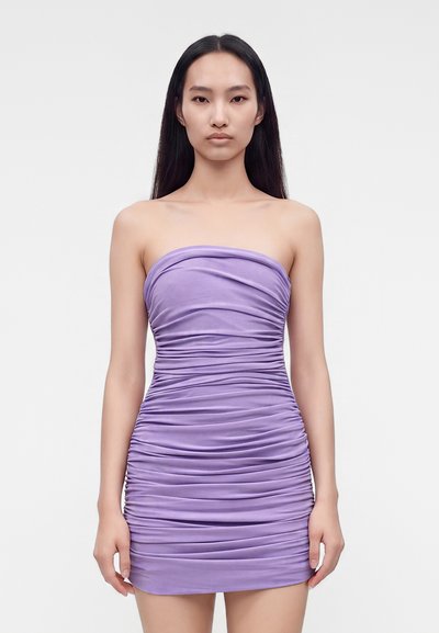 Kvinde med langt mørkt hår iført en strapless, rynket lilla minikjole, der står foran en ensfarvet lys baggrund.