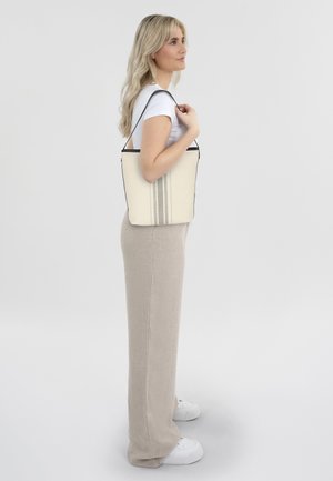 Canvas-Tote-Bag in Creme mit schwarzem Rand und drei vertikalen Streifen. Hat eine strukturierte Form und einen langen Schultergurt für einfachen Transport.