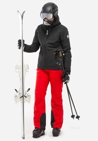 Skieur en veste noire, pantalon rouge, casque et lunettes de ski tenant des skis blancs et des bâtons de ski noirs sur un fond uni.